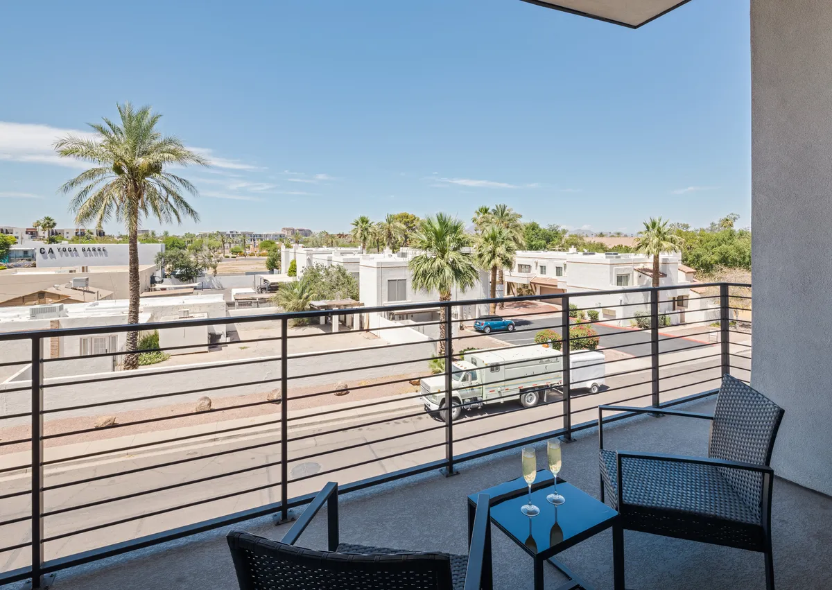 A view from a balcony furnished with comfortable seating, overlooking palm trees and a bustling street below. The atmosphere is calm, with sunset light casting a warm glow.