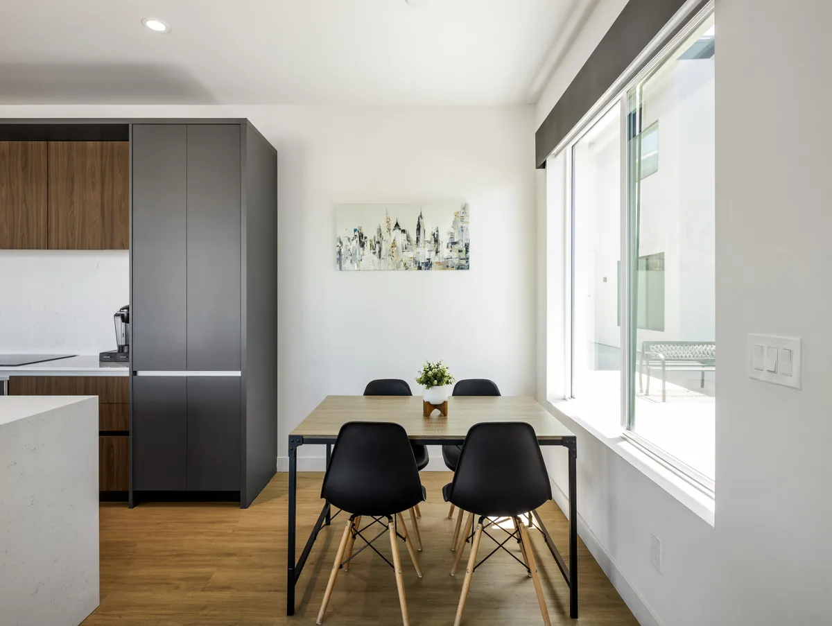 A cozy, contemporary dining area with a modern table set beneath warm lighting, adjacent to kitchen cabinetry and a window that lets in natural light. Art on the wall and thoughtful decor contribute to a welcoming ambiance for shared meals.