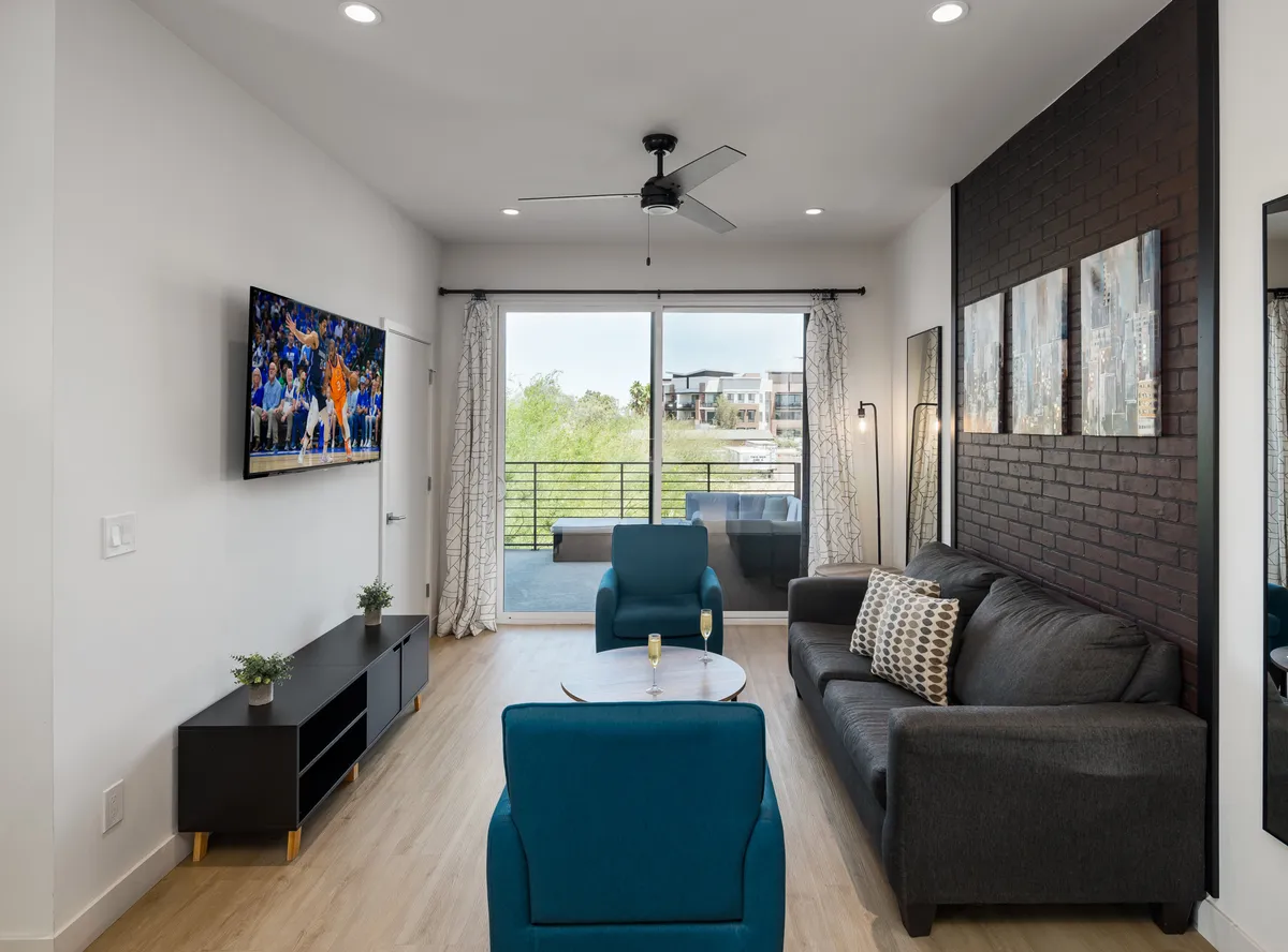 A sun-filled living room with a plush sofa faces a wall-mounted Smart TV, complemented by a sleek coffee table. Large glass doors open onto a balcony, allowing natural light to flood the space and offering views outdoors. The ambiance is casual and comfortable, perfect for relaxing or hosting friends.