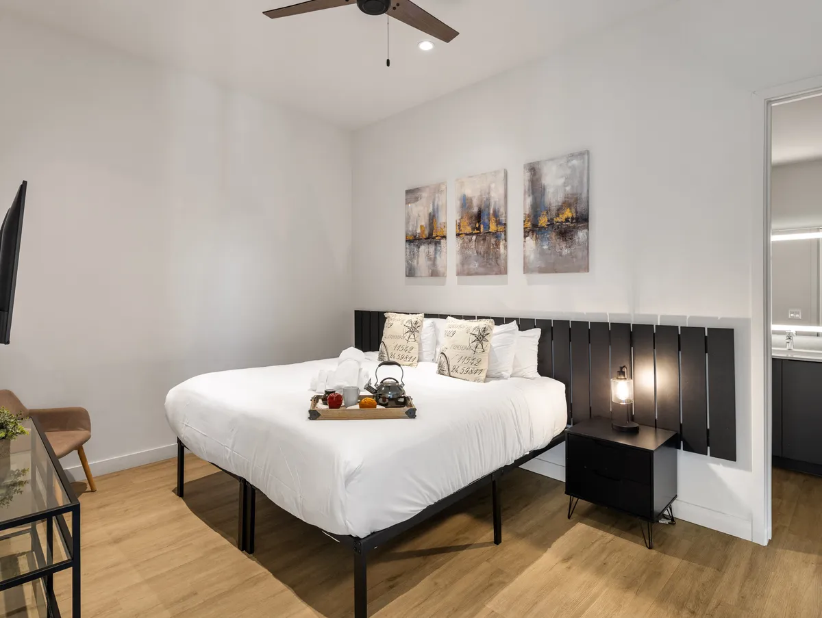 A cozy and stylish bedroom featuring decorative pillows on the bed, elegant art pieces on the wall, and a bedside lamp, evoking the feel of a hotel suite. Warm light and soft textures create a sense of luxurious comfort.