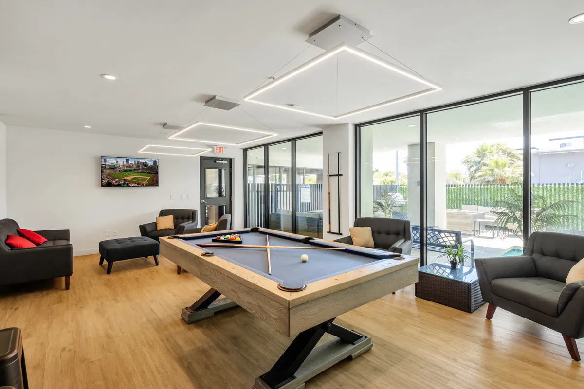 A vibrant communal clubhouse interior featuring a sleek pool table, comfortable sofas, a large television, and expansive windows. The space is well-lit and designed for both relaxation and friendly competition, inviting guests to unwind or socialize in style.