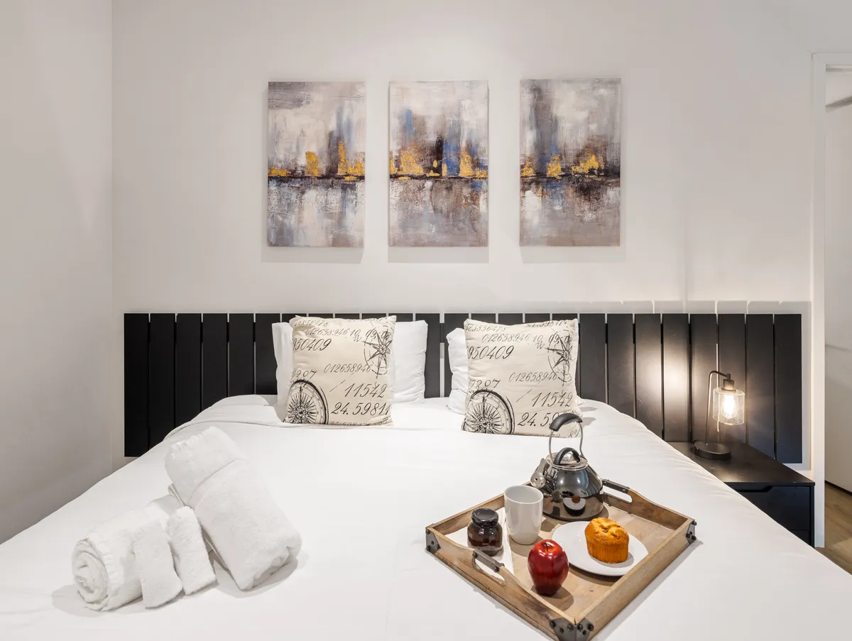 A serene bedroom with a neatly made bed adorned with decorative pillows, tasteful wall art, and a breakfast tray set for a cozy morning. The room is softly lit and imbued with a sense of relaxation and comfort.