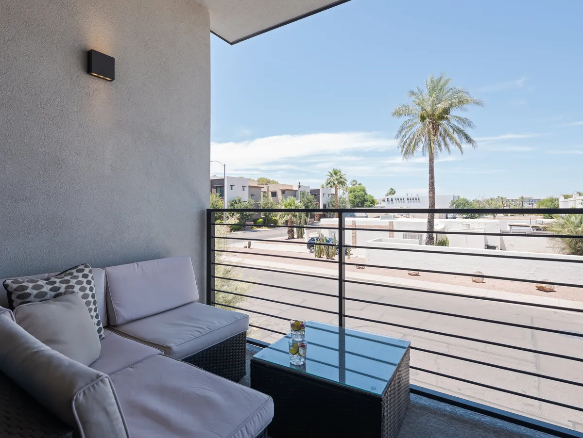 A private balcony with comfortable seating and a glass table overlooks a lively street framed by palm trees. The scene is inviting, with soft lighting suggesting an ideal spot to enjoy morning coffee, take calls, or simply relax while soaking up the city atmosphere.