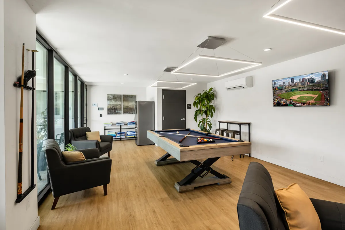 A vibrant clubhouse interior featuring a pool table, plush sofas, a television, and tasteful indoor plants. The space exudes a casual, fun atmosphere ideal for friendly competitions, watching games, or gathering for intimate group activities.