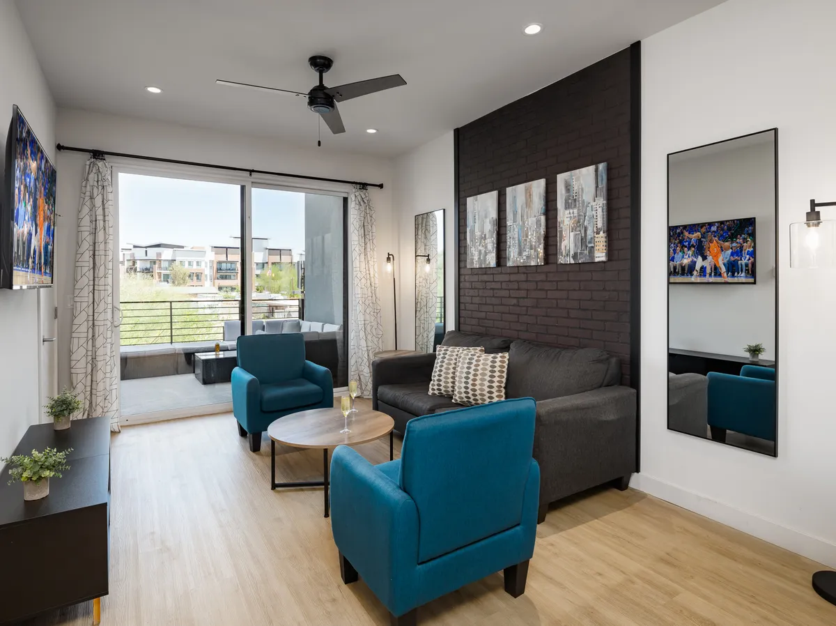 A contemporary living area featuring a comfortable sofa flanked by bold blue chairs, striking wall art, and large windows that invite sunlight into the space. The Smart TV and stylish decor make it ideal for both quiet mornings and lively evenings.